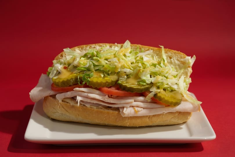 Turkey sub sandwich with lettuce, pickles, tomatoes, and sauce on a white plate against a red background.