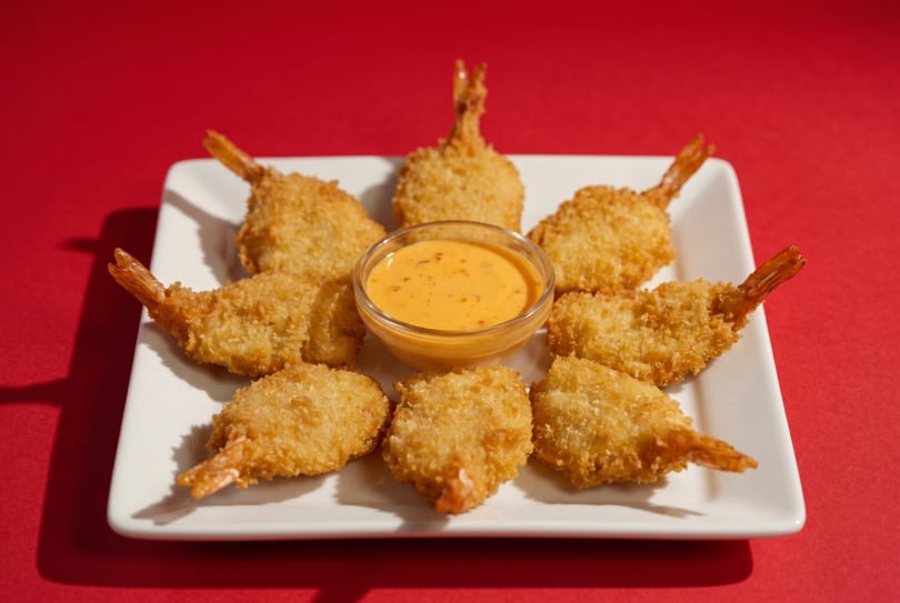 Eight golden-brown fried shrimp with tails on, arranged around a bowl of orange dipping sauce on a white plate.