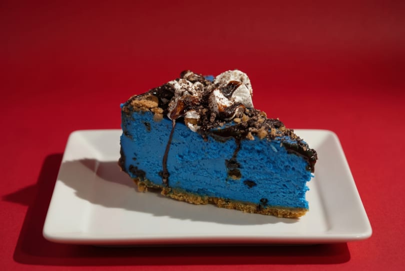 Slice of vibrant blue cheesecake with whipped cream, chocolate drizzle, and cookie crumbles on a white plate.