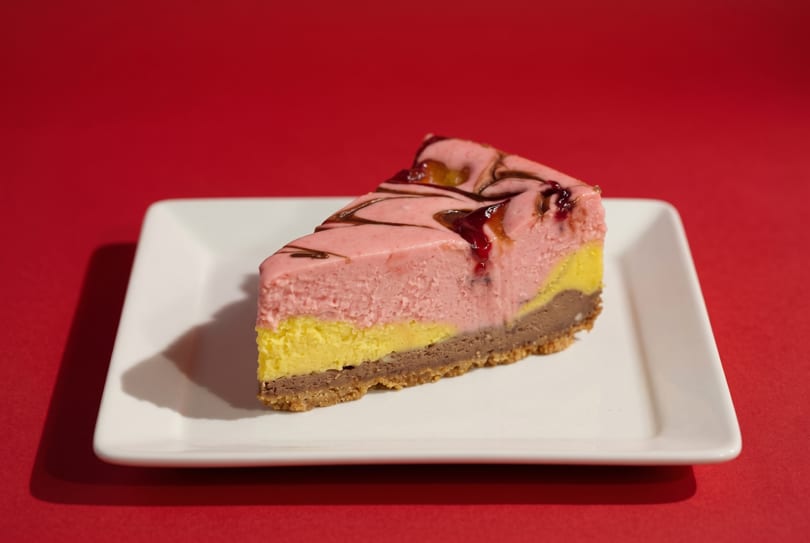 Slice of layered pink, yellow, and brown cheesecake with chocolate and berry drizzle on a white plate.